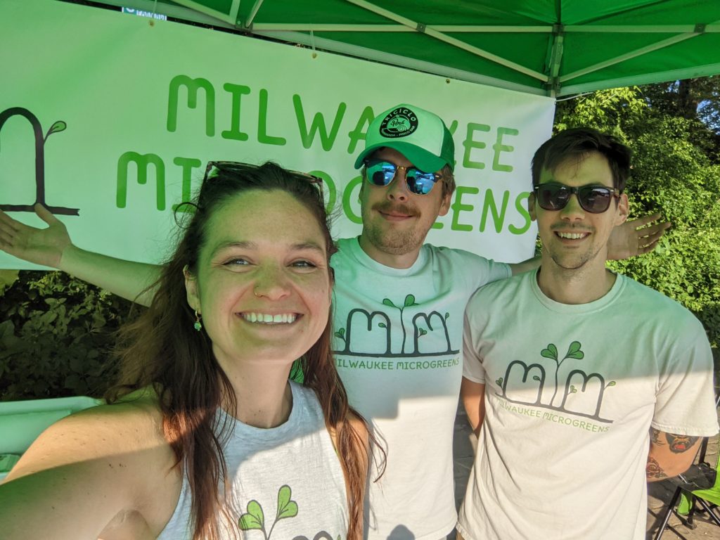 Selfie of the owners of Milwaukee Farmers United and Milwaukee Microgreens wearing their own merch beaming at the camera at a farmers market.